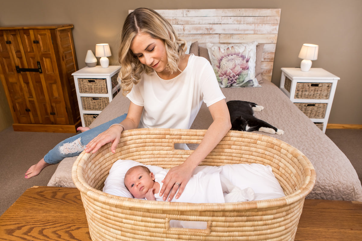beige bedroom scene with blonde haired lady looking over her newborn baby in a grass crib
